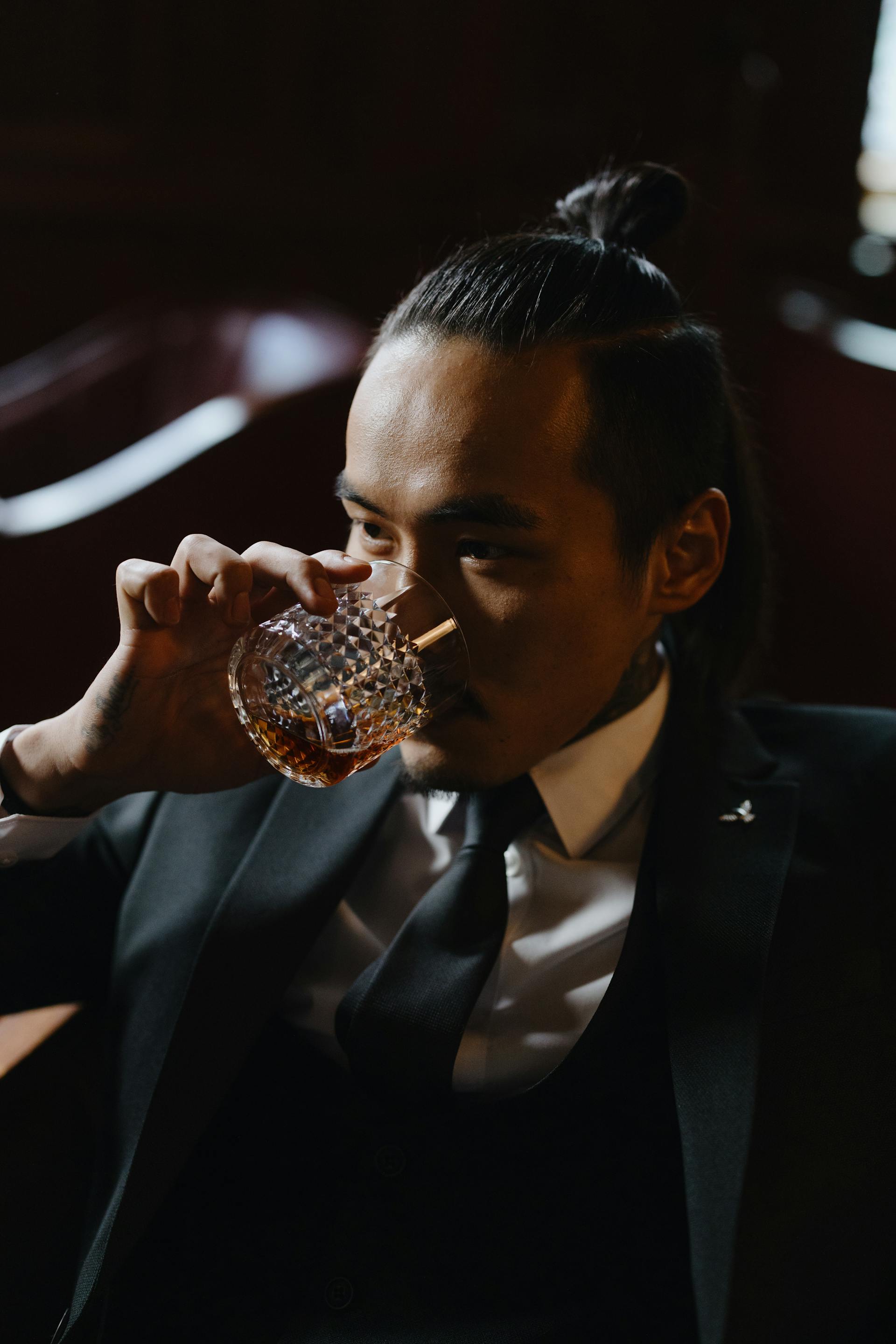 A close-up of a man with a top knot and black suit taking a sip of amber-colored liquid from a crystal tumbler in a dark, moody setting.