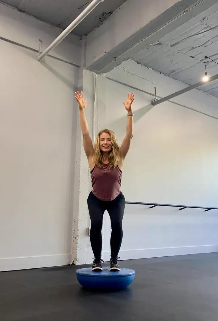 Heather Hausman with long blonde hair stands on a blue balance trainer with her arms raised, set against a spacious industrial-style fitness studio backdrop featuring white walls and visible ceiling pipes.