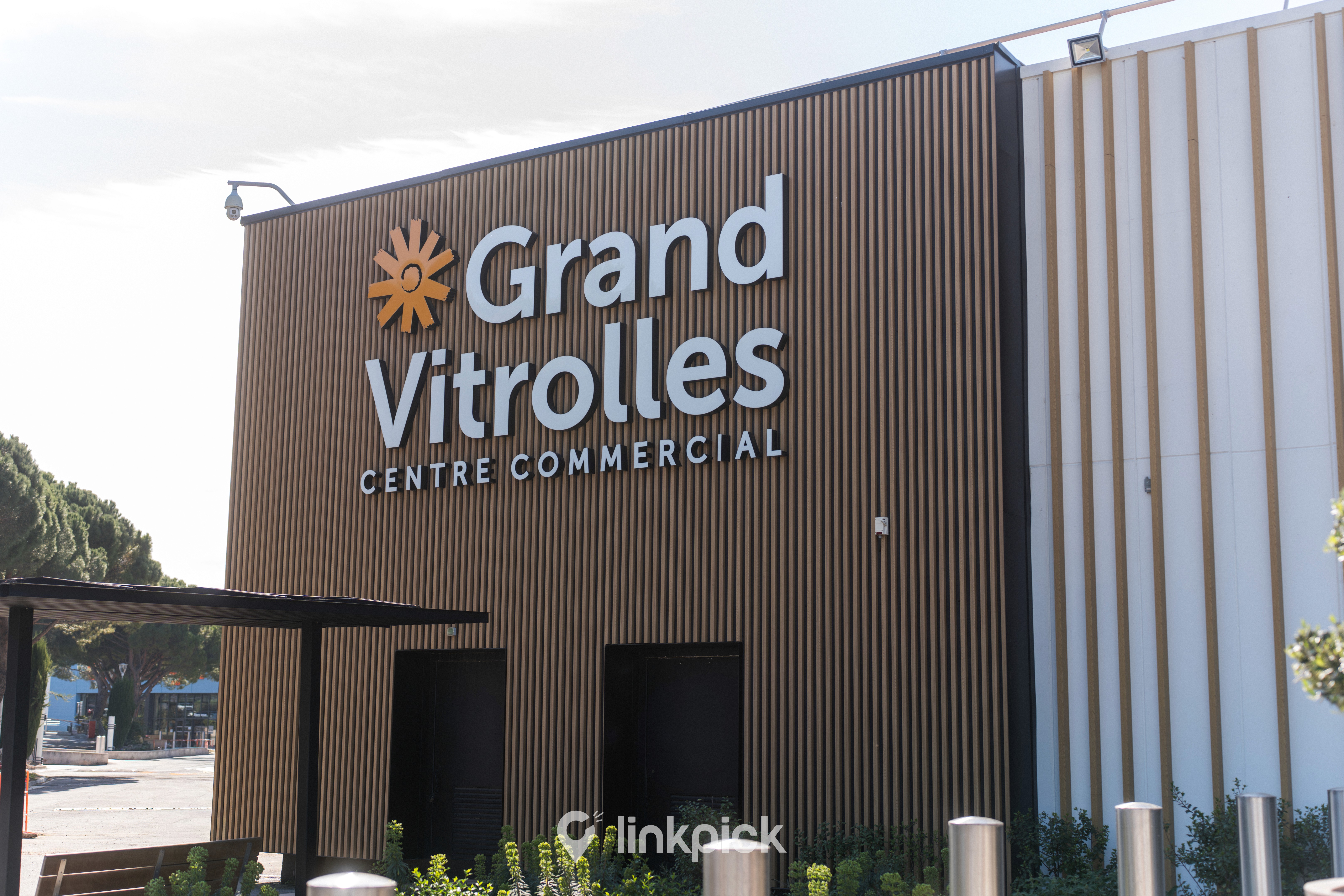 Vue extérieure détaillée de l'entrée principale du Centre Commercial Grand Vitrolles par une journée ensoleillée. La façade est habillée d'un revêtement moderne en tasseaux de bois marron texturé. En haut à gauche, un grand logo orange en forme de soleil estilé est visible. Juste à côté, l'enseigne 'Grand Vitrolles' est écrite en grandes lettres blanches sans-serif tridimensionnelles. En dessous, 'CENTRE COMMERCIAL' est inscrit en plus petites lettres blanches similaires. L'entrée comporte deux portes sombres encastrées. Au premier plan, des plantes vertes d'aménagement paysager et des poteaux en acier inoxydable complètent la scène. Le logo 'linkpick' est subtilement visible dans le coin inférieur droit parmi la végétation.