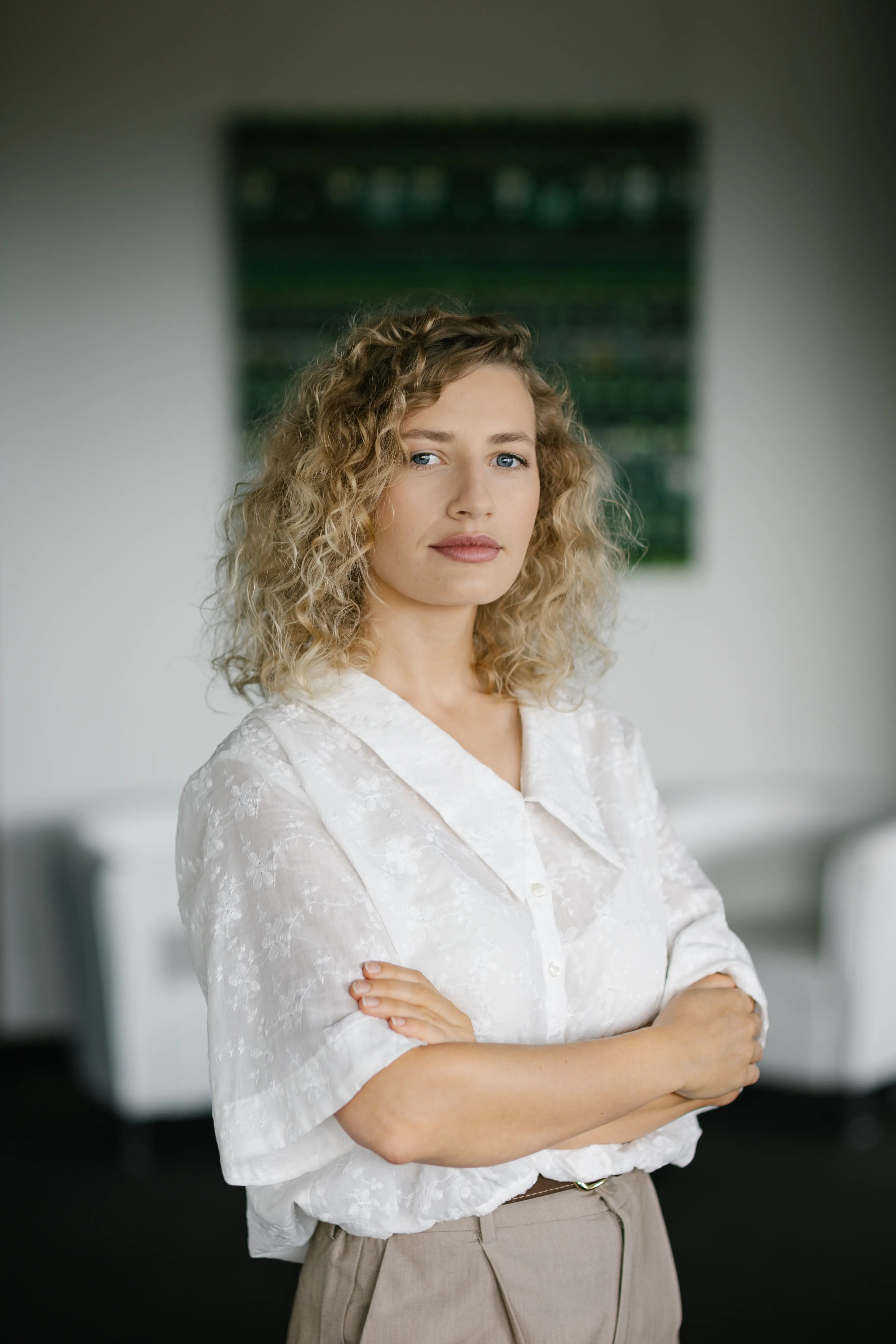 Kobieta z kręconymi blond włosami ubrana w białą bluzkę stoi ze skrzyżowanymi ramionami, patrząc pewnie w kamerę. Tło jest delikatnie rozmyte, z białymi meblami i ścianą z zieloną grafiką.