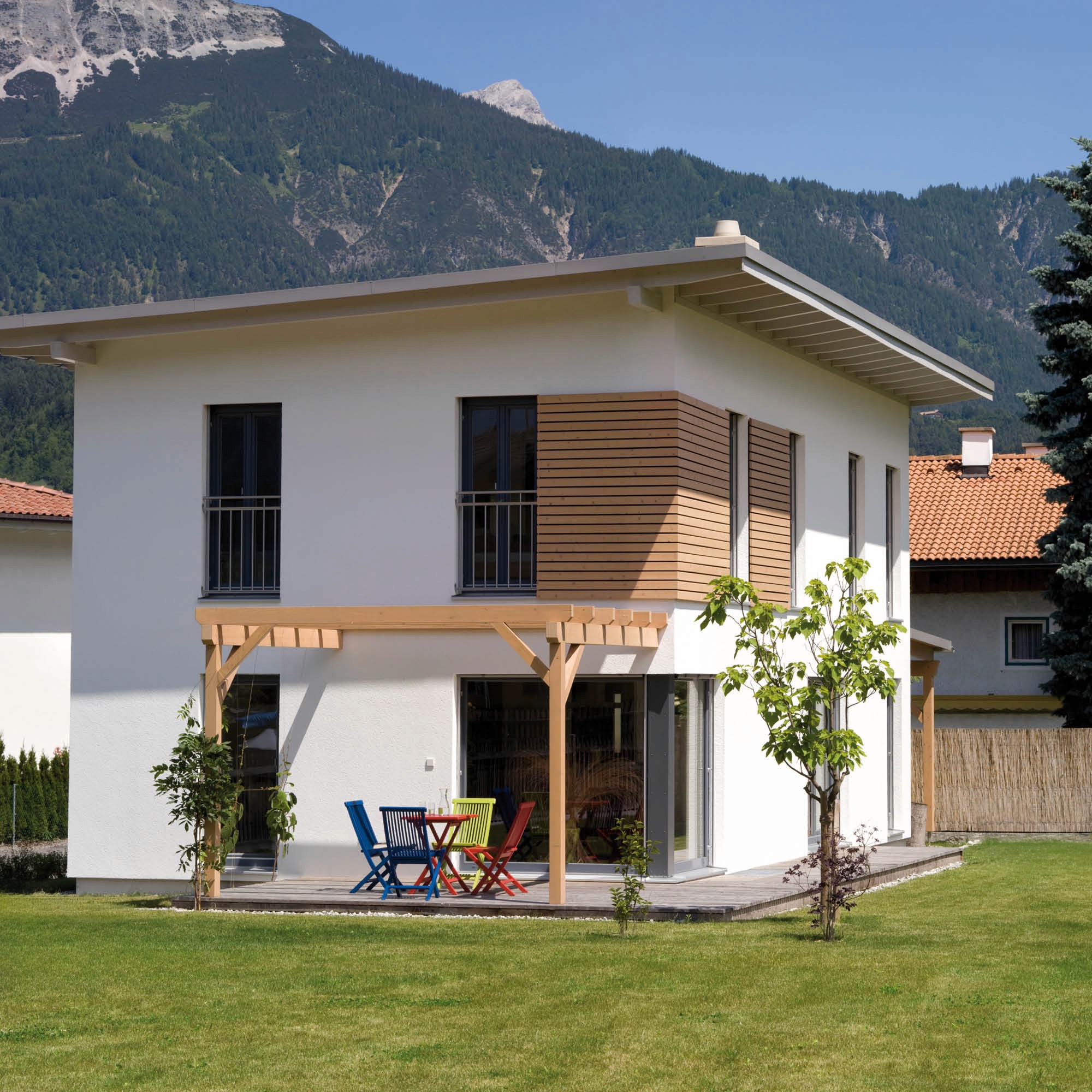Modernes weißes Haus mit Holzverkleidung, Terrasse und Bergblick, auf der Terrasse bunte Stühle und ein Tisch.