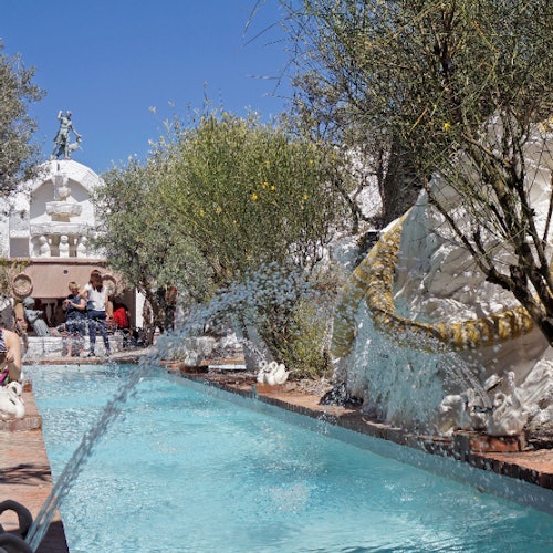 Outdoor scene with a pool, statues, spraying water, trees, and people sitting in the background under a clear blue sky.
