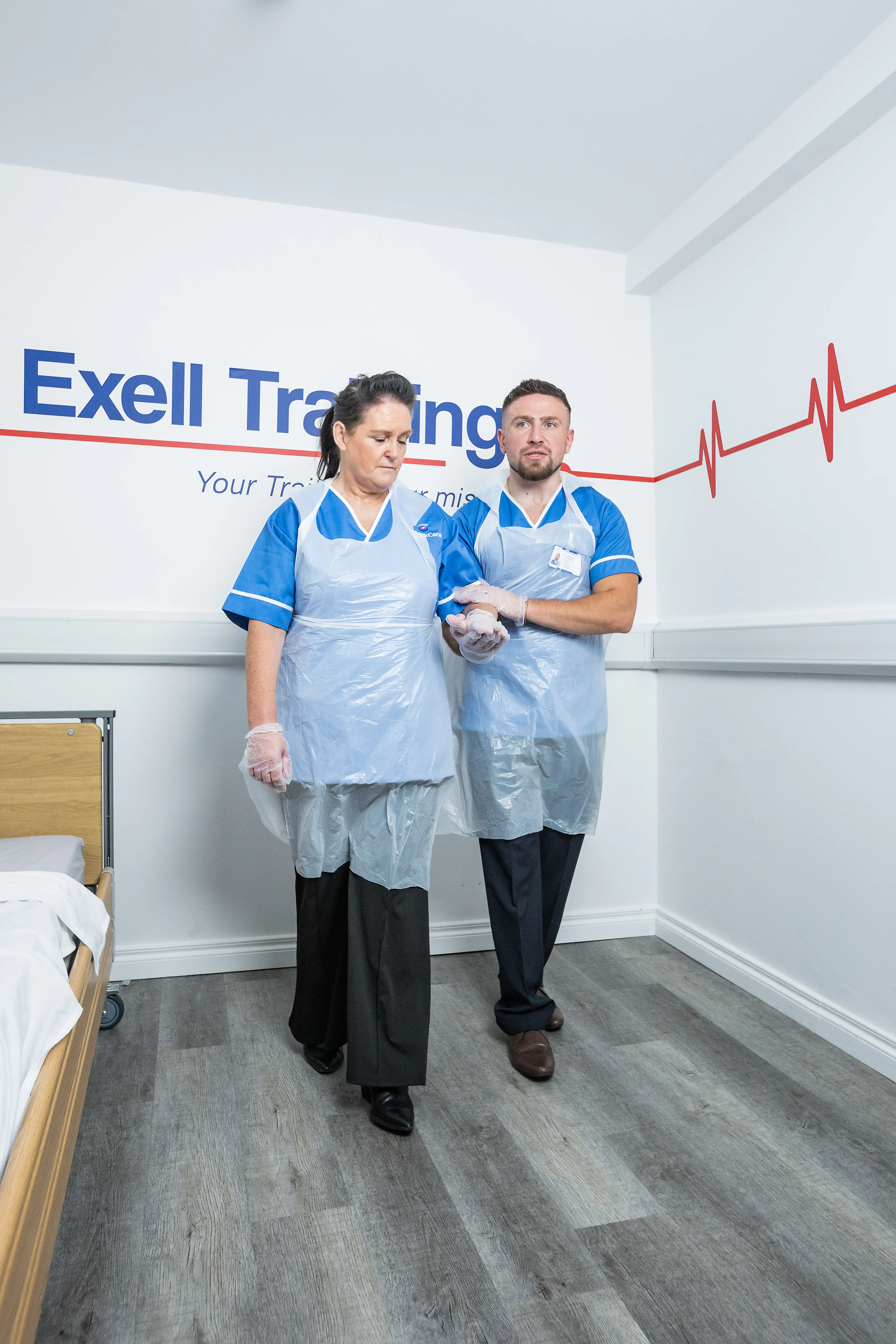 Two healthcare professionals in training uniforms and aprons practising a patient support technique in a clinical training room with Exell Training branding on the wall.
