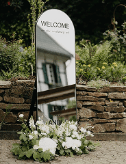 Willkommensschild auf Spiegel mit floraler Dekoration für eine Hochzeit im Außenbereich