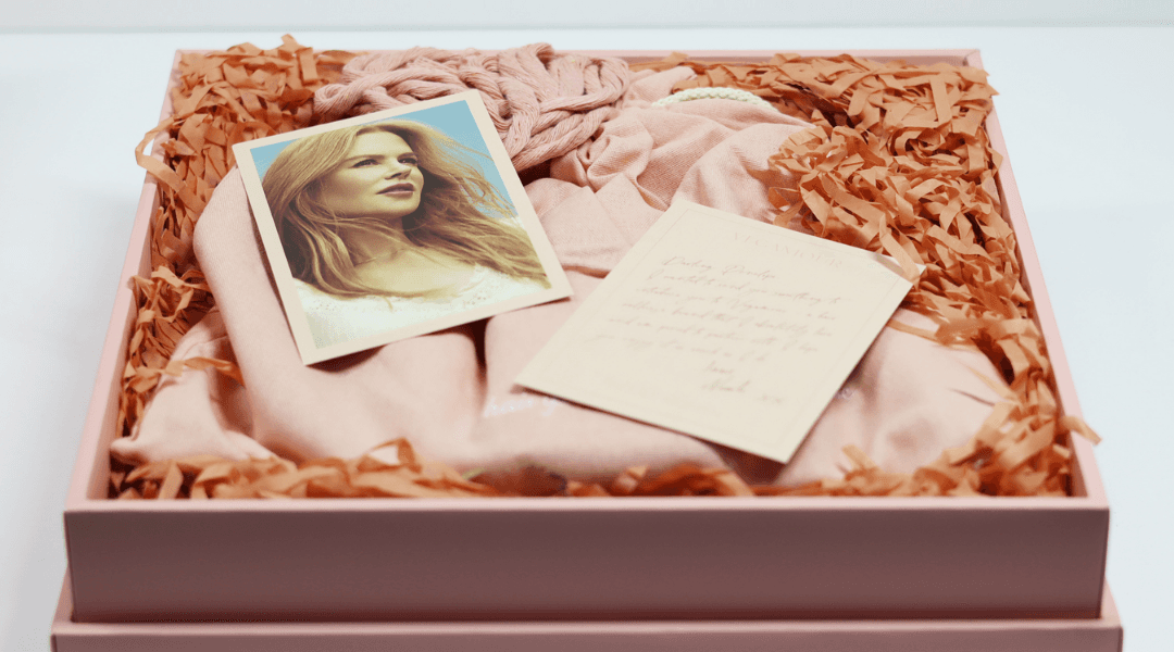 A blush pink box with pink crinkle paper, a note, the merch item, and a picture. The box is open and on a white table.