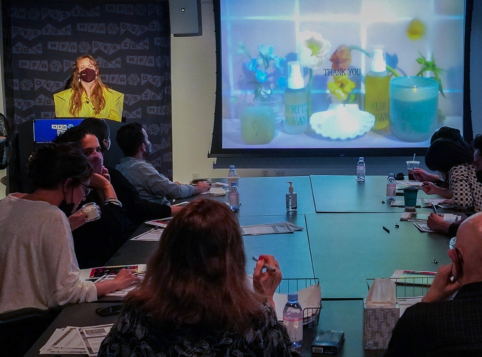 A woman dressed in yellow is giving a speech in front of people sitting around a table, and there is an image projected on the right side of the wall with miscellaneous objects near the wall