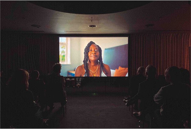 Audience seated in a dark room watching a large screen showing a woman carer speaking in a brightly lit room, during a film presentation.