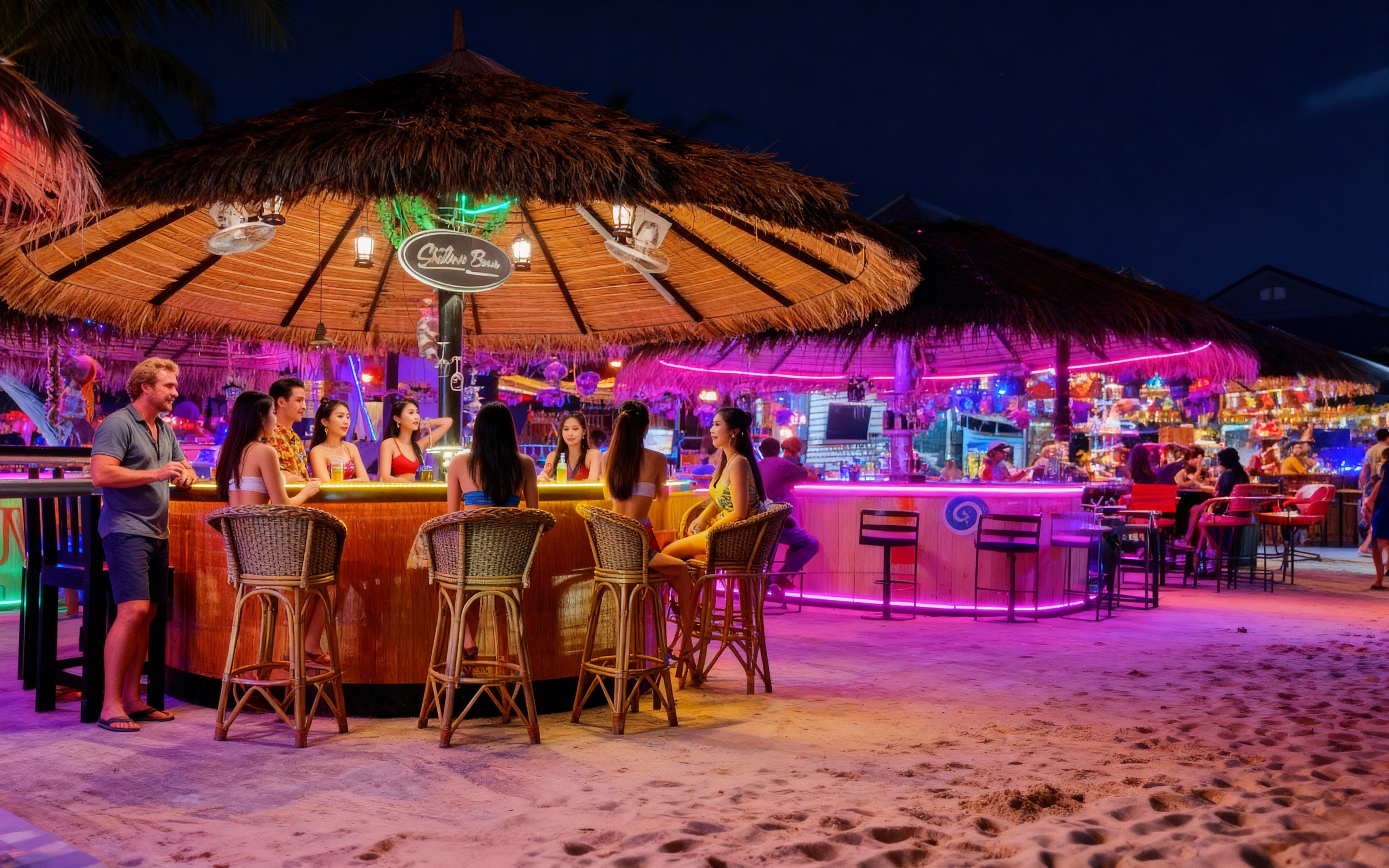 Nighttime scene at Wonderland Bar in Hua Hin with groups of people chatting at a thatched-roof bar lit by colorful neon lights, surrounded by beach sand, palms, and adjacent tiki huts.