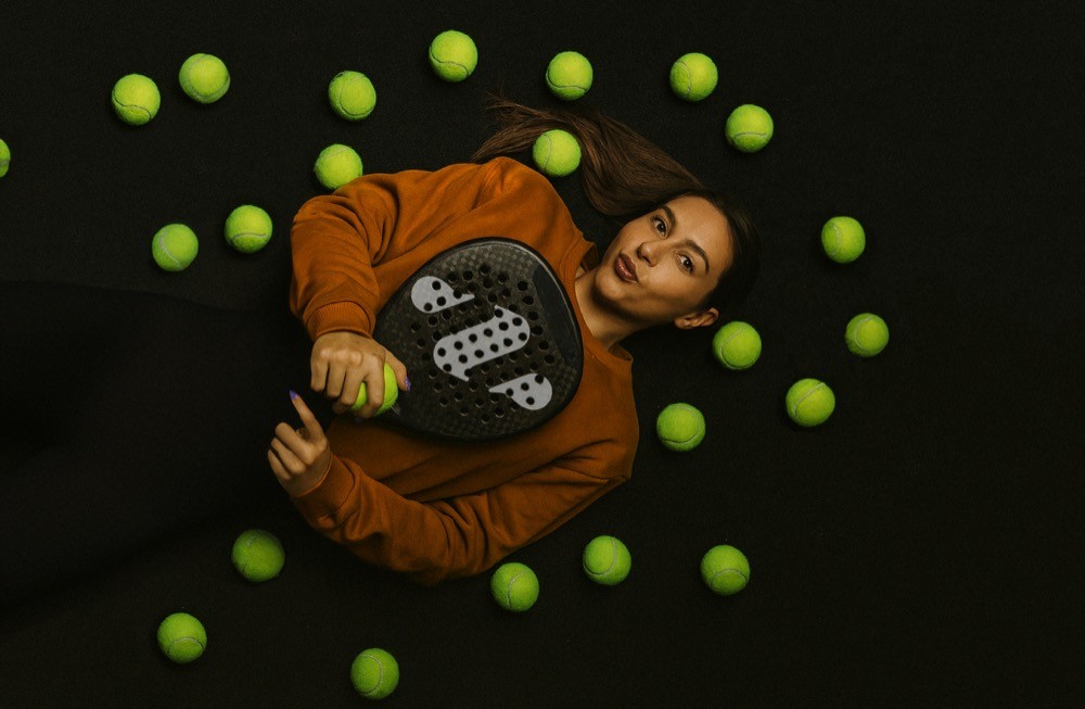 Padelspielerin liegt auf dem schwarzen Court, umgeben von gelben Bällen – ein kreativer Moment zwischen Sport und Stil bei Padelnero.