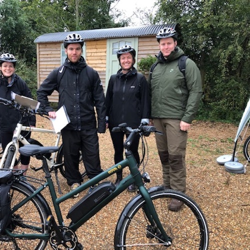 Four people with helmets stand next to bicycles in front of a small wooden hut and trees.