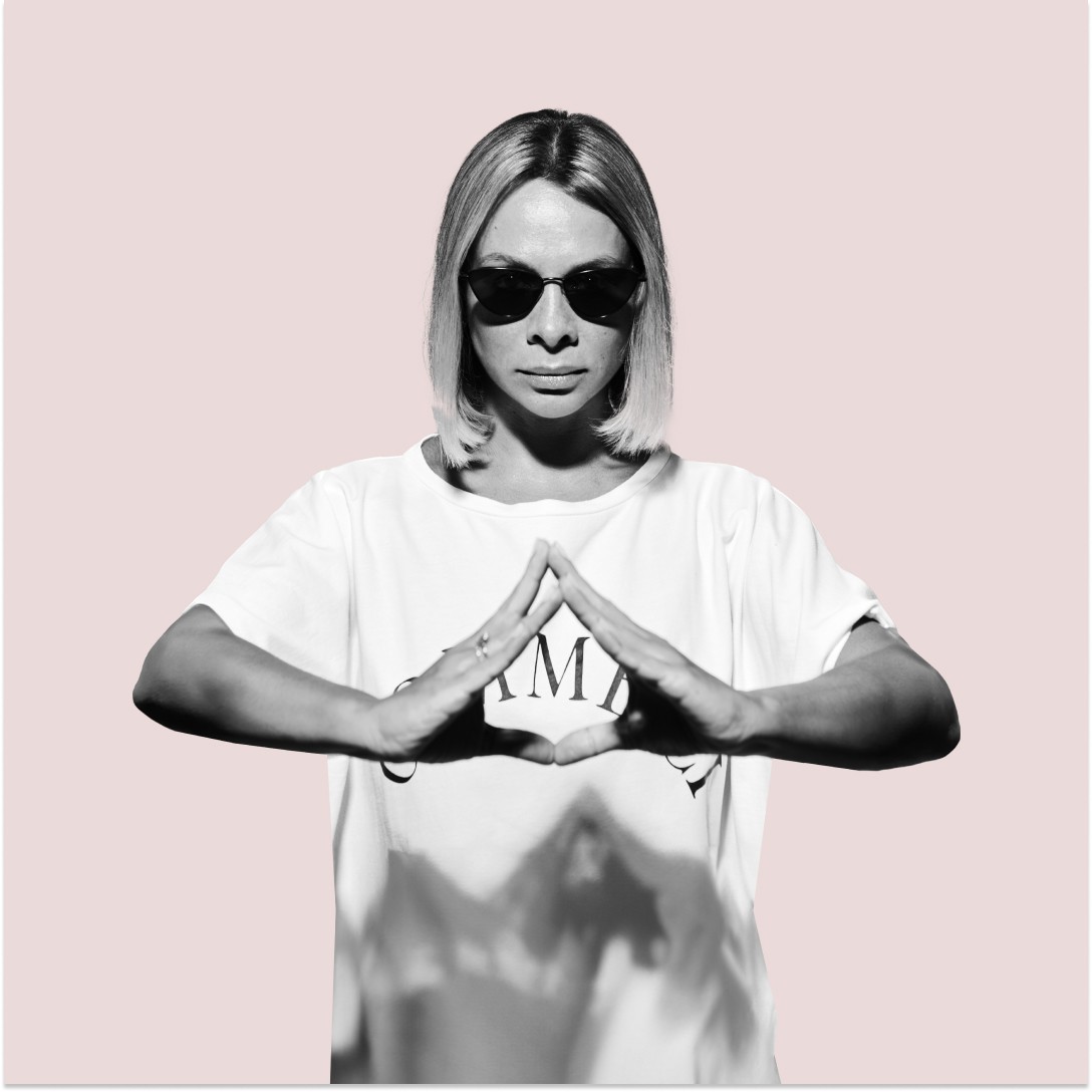 Smiling blonde woman with freckles in a white shirt, arms crossed, minimalist studio portrait.
