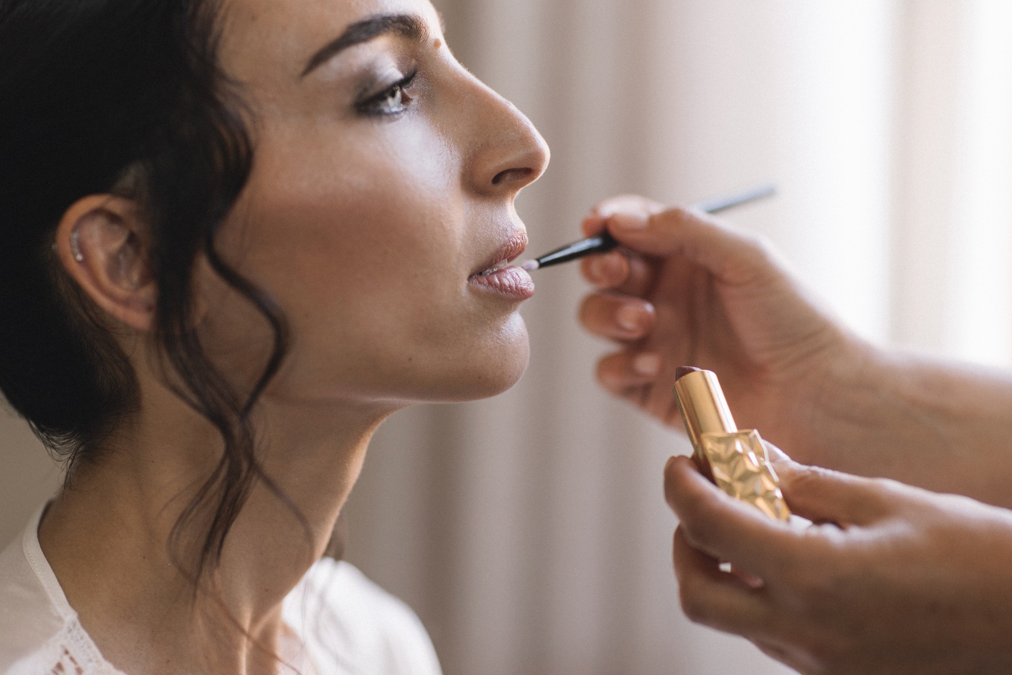 Sposa ai preparativi matrimoniali: trucco labbra durante i preparativi, luce naturale dai tendaggi bianchi