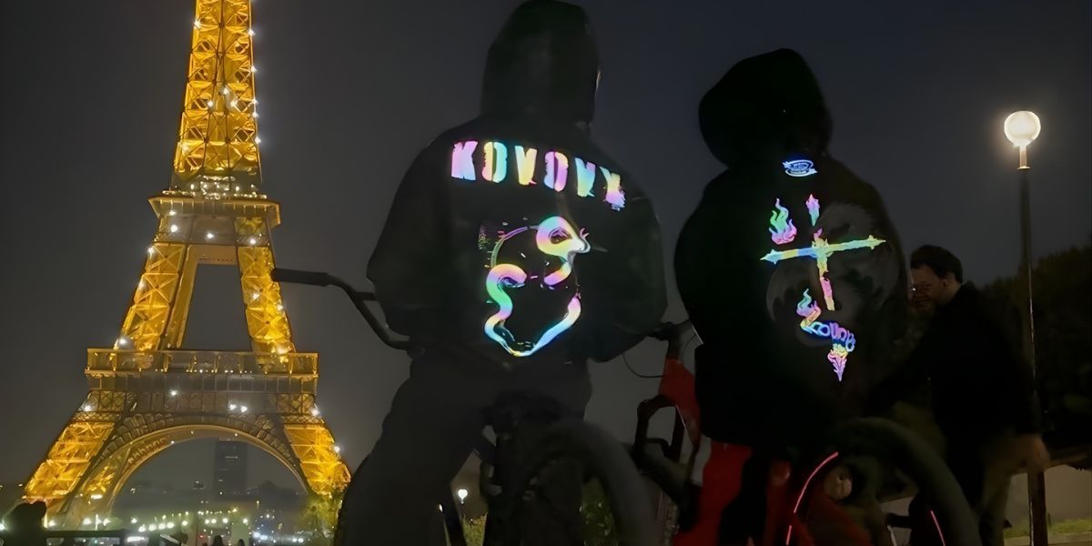 Two people in Kovovy glow-in-the-dark hoodies with reflective designs biking in front of the illuminated Eiffel Tower.