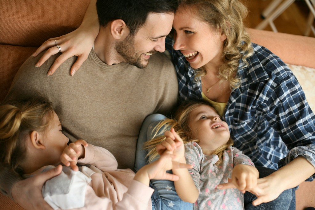 A joyful family cuddling and laughing together on a couch, with parents embracing their two young daughters, creating a warm and loving atmosphere.