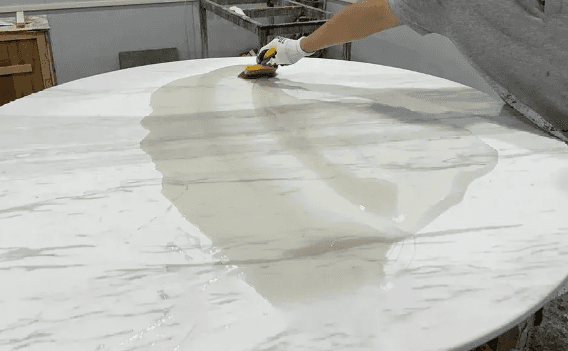 A person applying a liquid treatment to a large, round marble tabletop with a smooth, white surface and gray veining. The scene is inside a stone workshop.