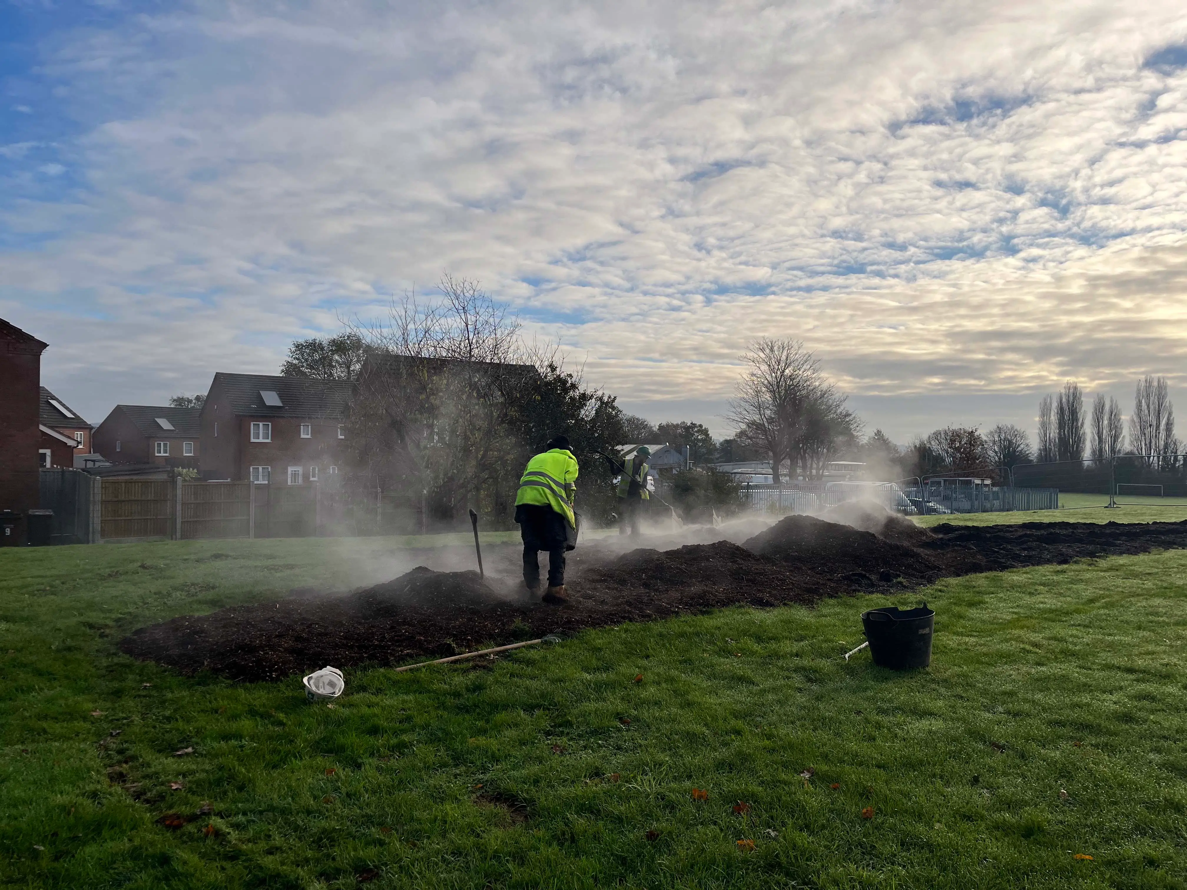 A worker sprays water on a field under a cloudy sky, with mist rising and green grass in the foreground.
