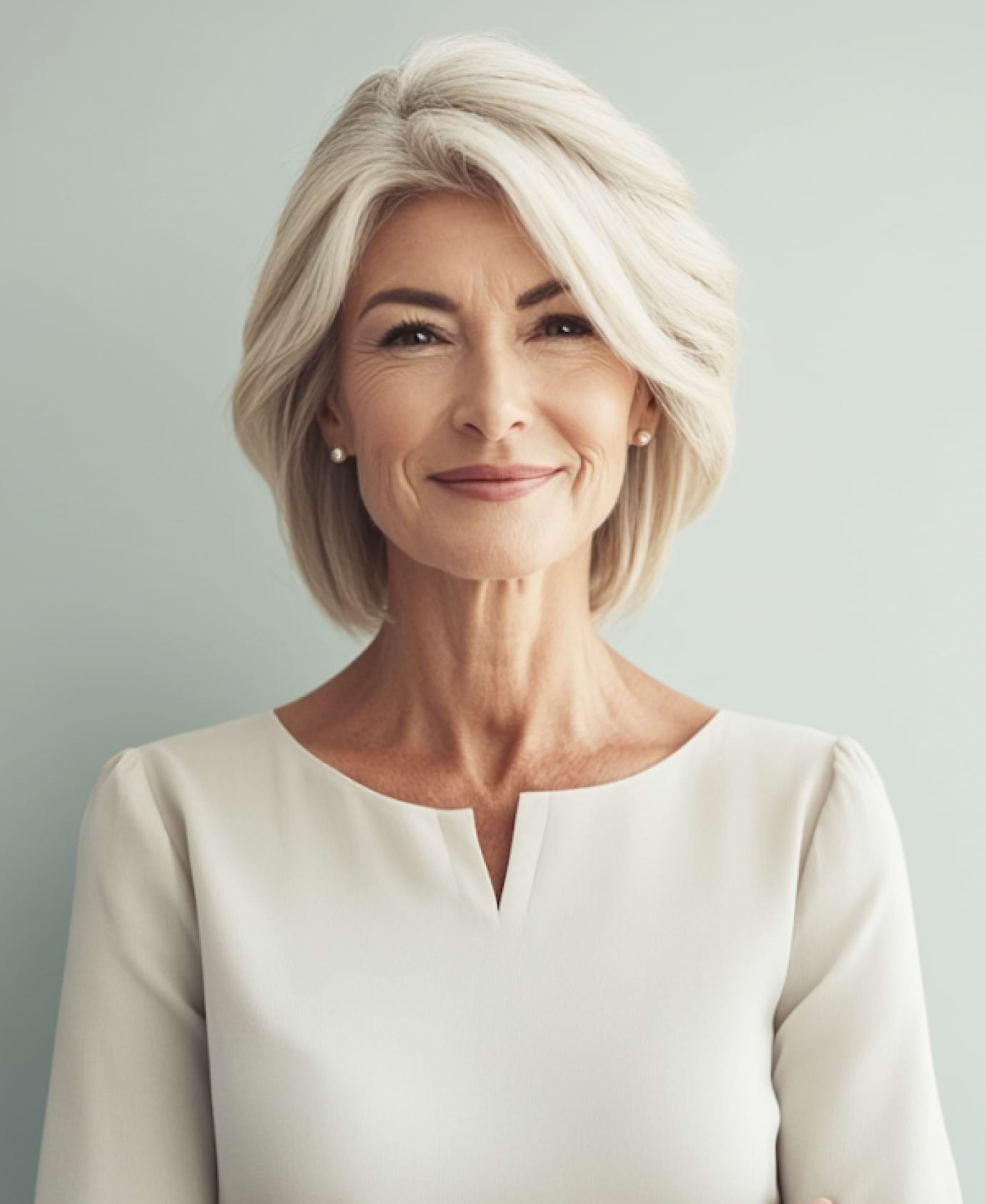 A confident woman with short white hair smiles gently, wearing a light-colored blouse against a soft, pale background, conveying warmth and professionalism.