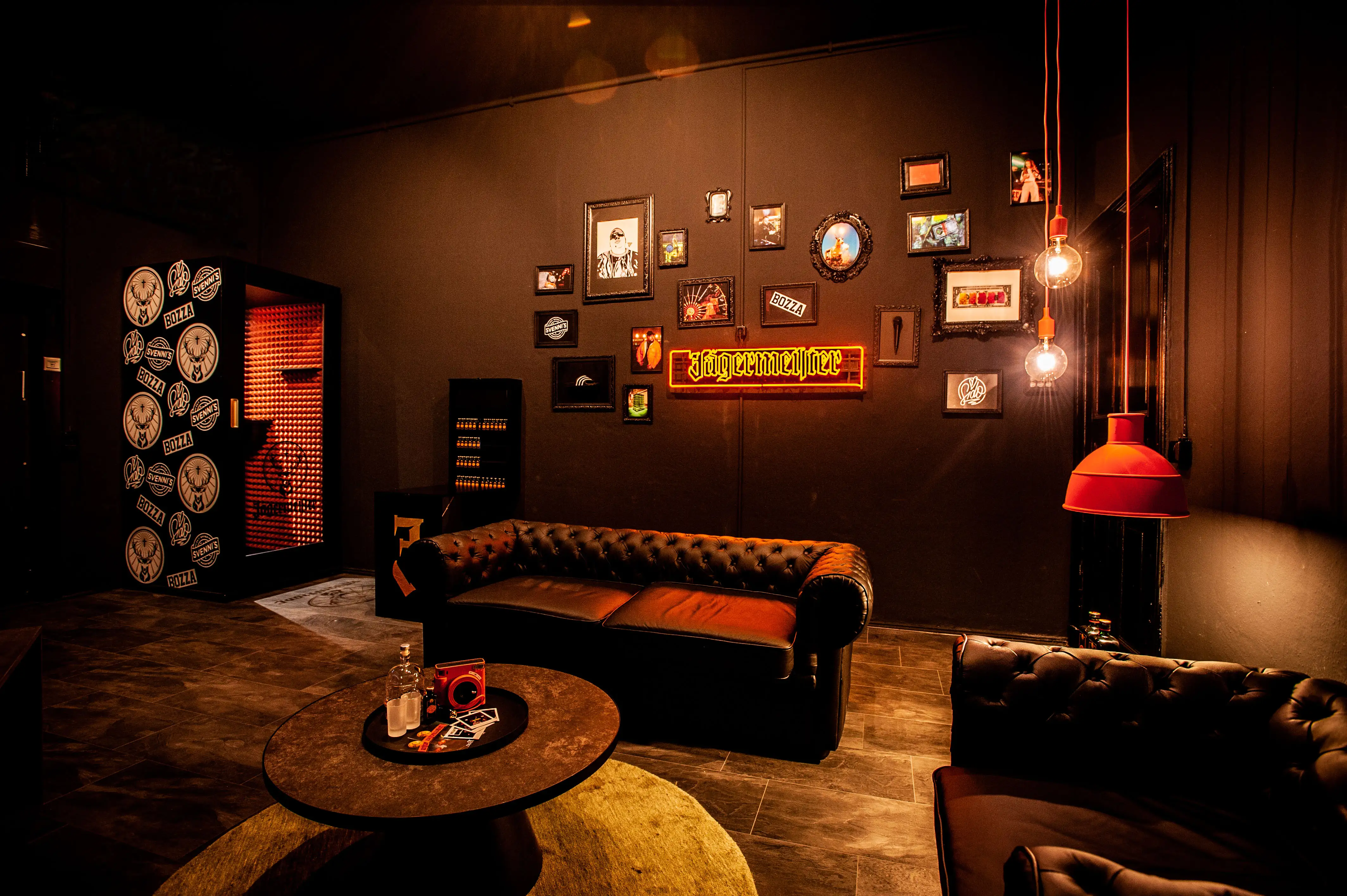 Lounge at JÄGERMEISTER x SVENNI'S with leather Chesterfield sofas, a gallery wall of framed pictures, an orange Jägermeister neon sign, red pendant lamp and a door covered in coasters.