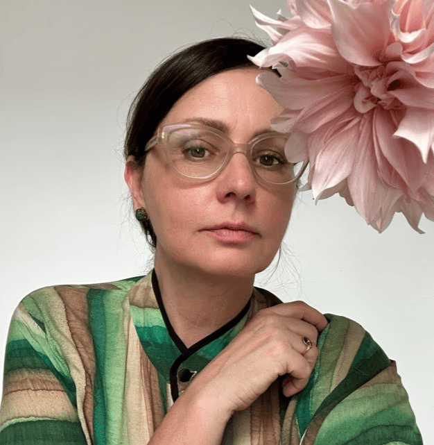 Retrato de Irina Khatsernova con gafas transparentes y blusa verde, acompañada por una flor rosada grande en primer plano.
