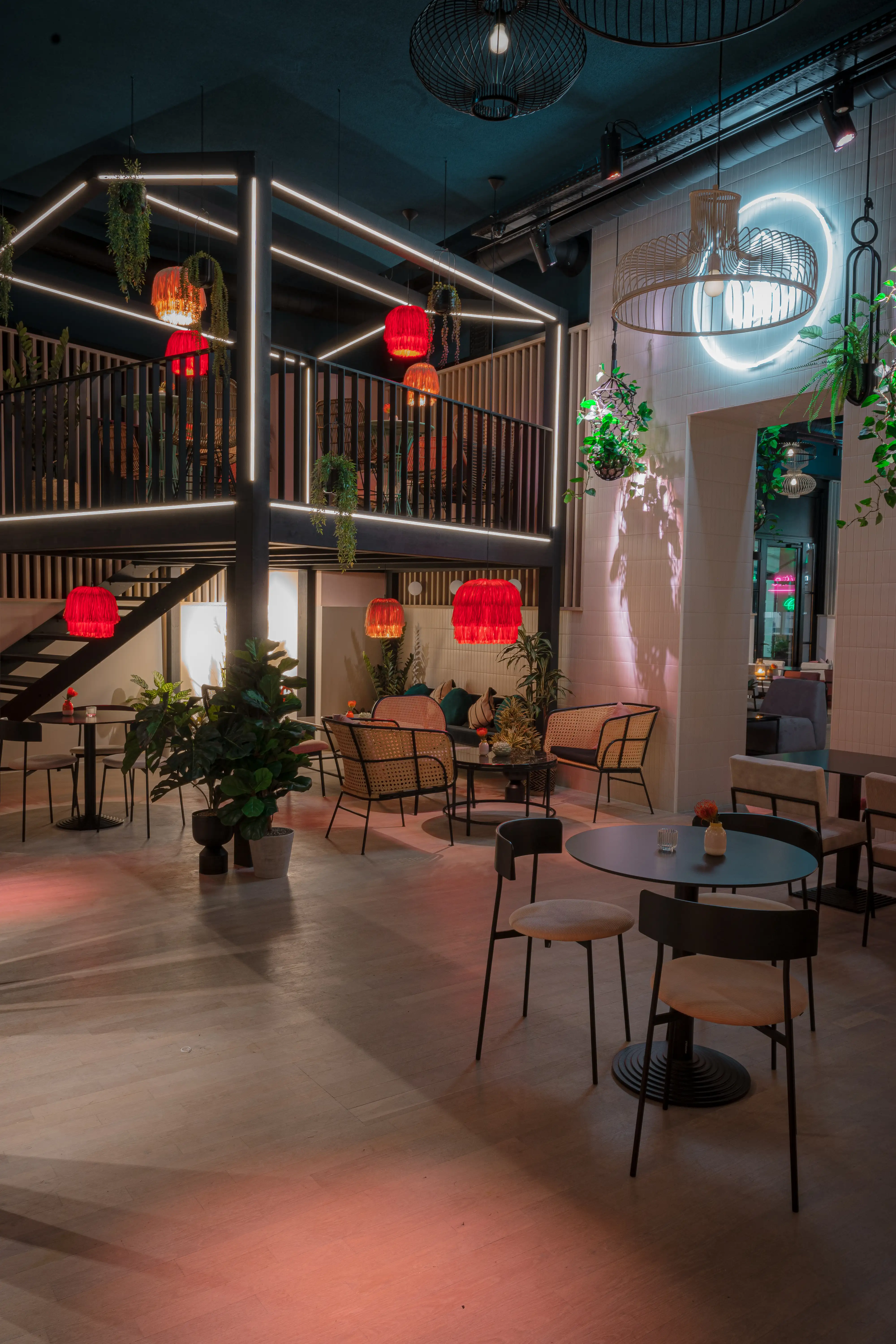 Evening view of LOKAL VIER with an illuminated black steel gallery structure, red fringe pendant lamps, rattan chairs, plants and small black bistro tables.