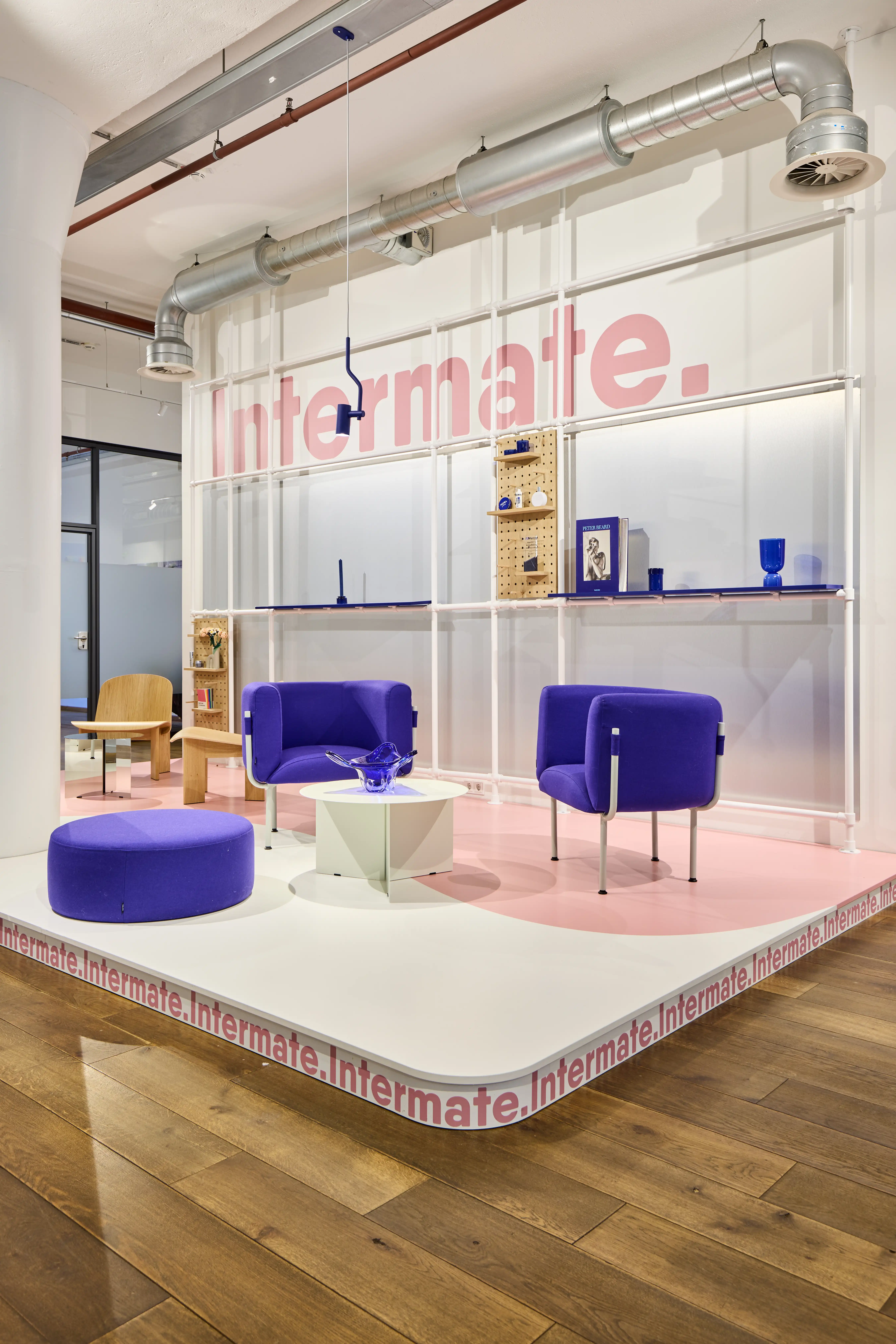 Lounge stage at INTERMATE OFFICE HAMBURG with purple armchairs and a purple pouf, a faceted white side table holding a blue glass bowl, pink Intermate lettering on a translucent shelf wall, and exposed silver ventilation ducts.