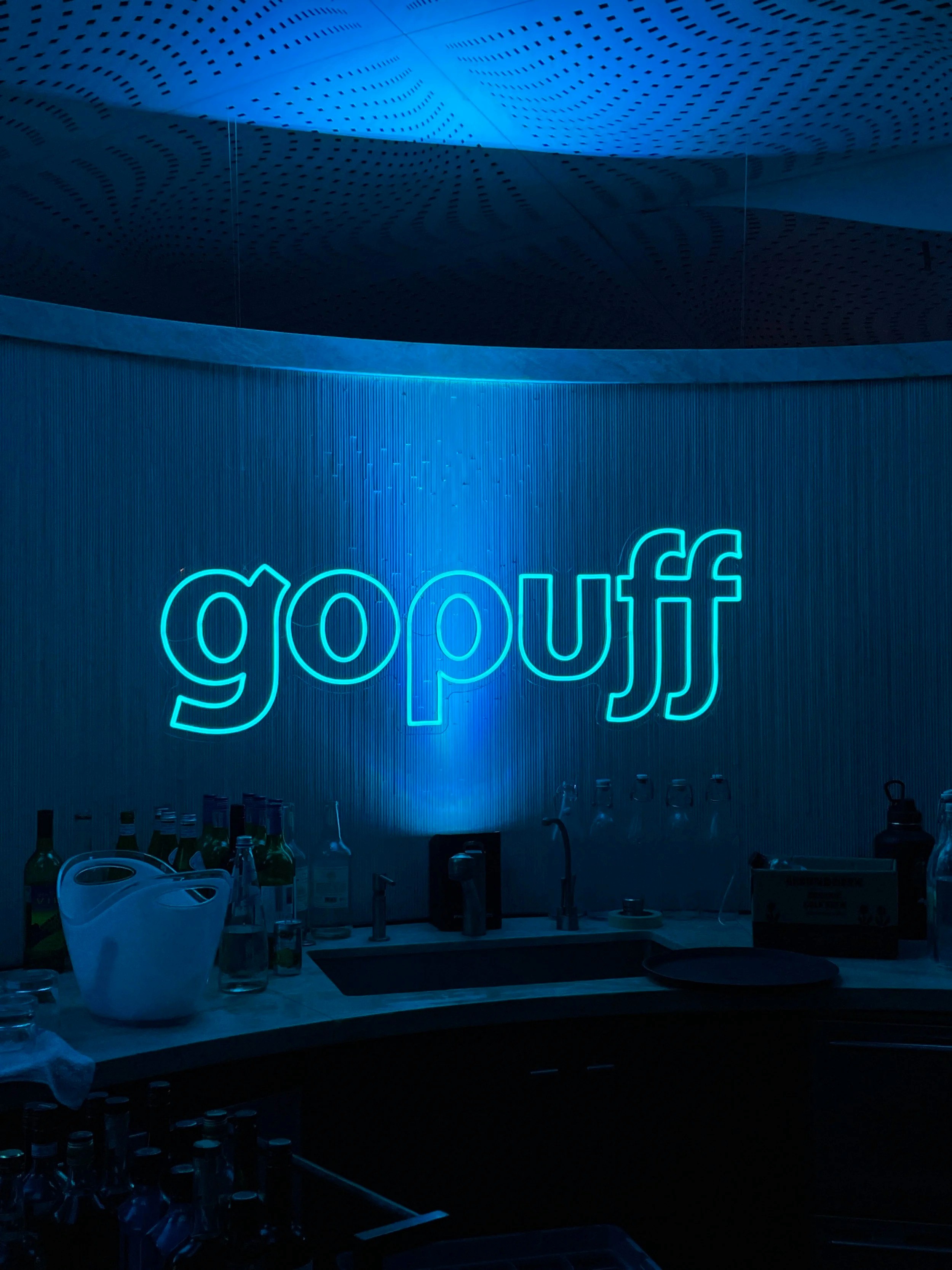 Large "gopuff" neon sign in cyan blue glowing against a textured wall behind a bar setup, illuminating the space with a blue glow from above.