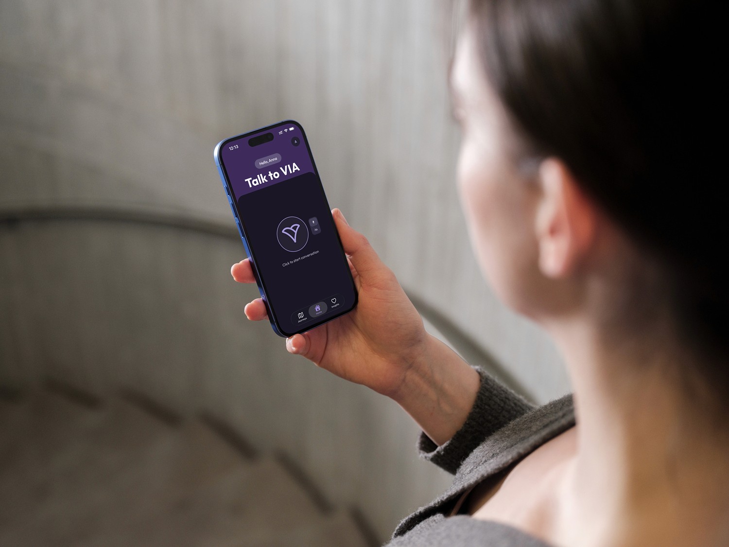 Over-the-shoulder shot of a woman standing in a modern concrete stairwell, looking at her smartphone. The screen displays the Vialog home interface with the "Talk to VIA" prompt and central interaction button, suggesting a moment of reflection during the day.