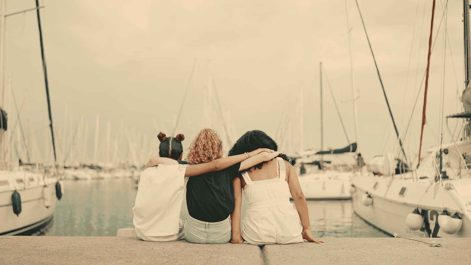 Three people sit on a dock with their arms around each other, facing a marina filled with white sailboats under a cloudy sky.