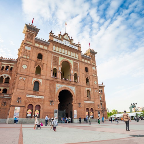 Duży ceglany budynek z łukowatym wejściem oznaczonym jako "Plaza de Toros" pod niebieskim niebem z rozproszonymi chmurkami, ludzie stojący i spacerujący na zewnątrz.