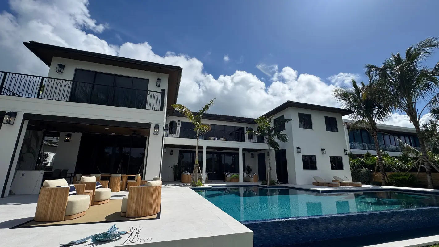 Rear elevation of a Bay Point waterfront estate, with a two-story facade, covered outdoor living area, lounge seating, palm trees, and a pool under a bright blue sky.