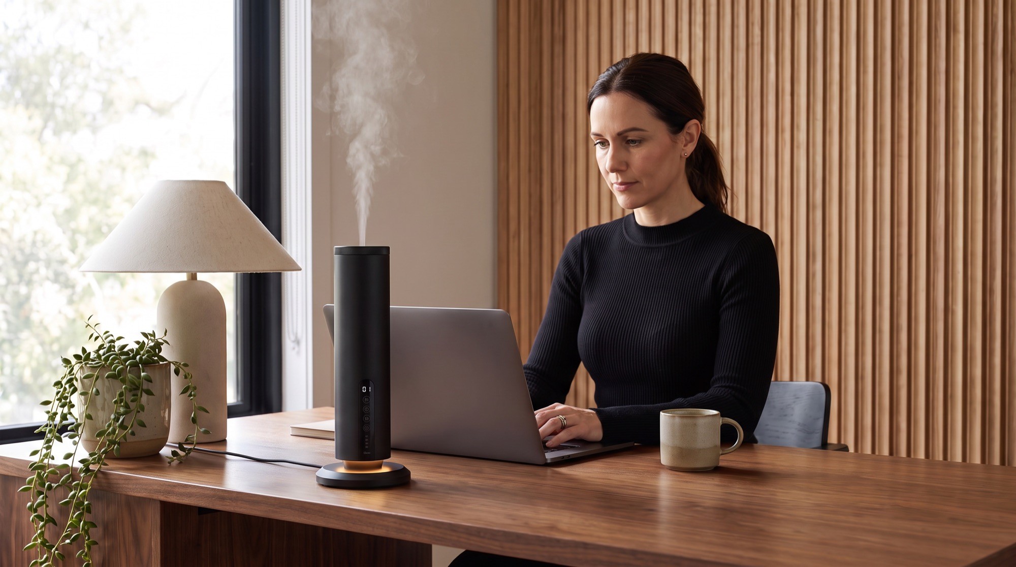 Woman working at a walnut desk beside a Scent Aura waterless diffuser releasing a fine, dry mist