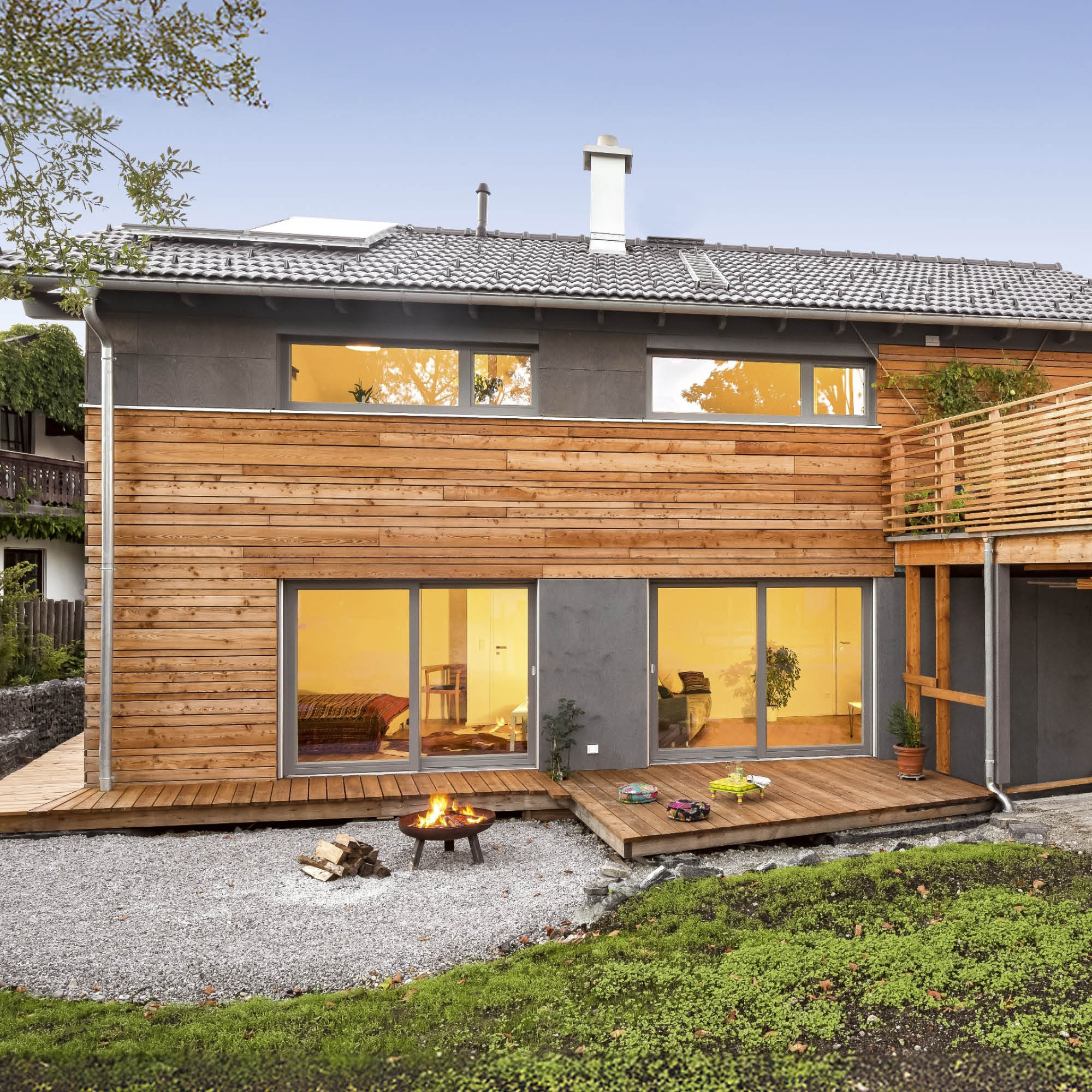 Modernes Holzhaus mit großer Glasfront beleuchteten Innenräumen, einem Kaminfeuer und Terrassenflächen.