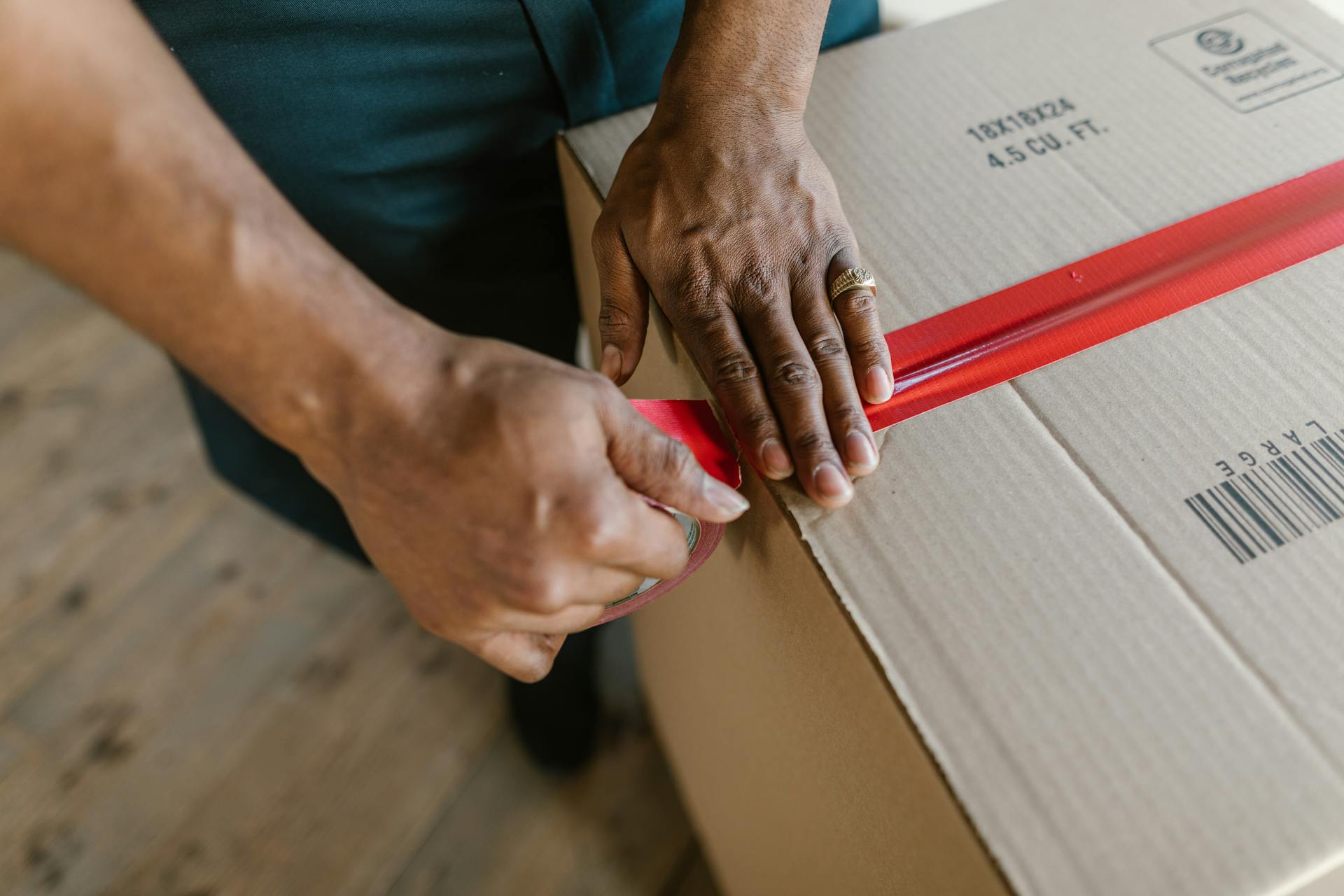 A close-up shot of hands using clear packing tape with a red filament to seal a large cardboard box.