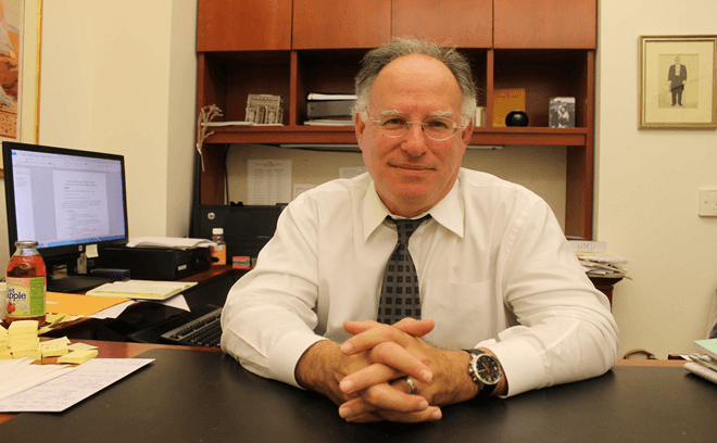 Judge Harry Dorfman's seated at a desk in an office, hands clasped, with shelves and documents behind him.
