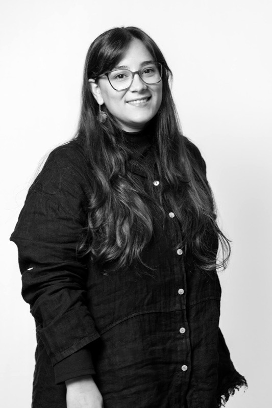 Black-and-white studio portrait of a smiling woman with long wavy dark hair, wearing glasses and dangling earrings with a black button-front linen shirt, against a plain light background.