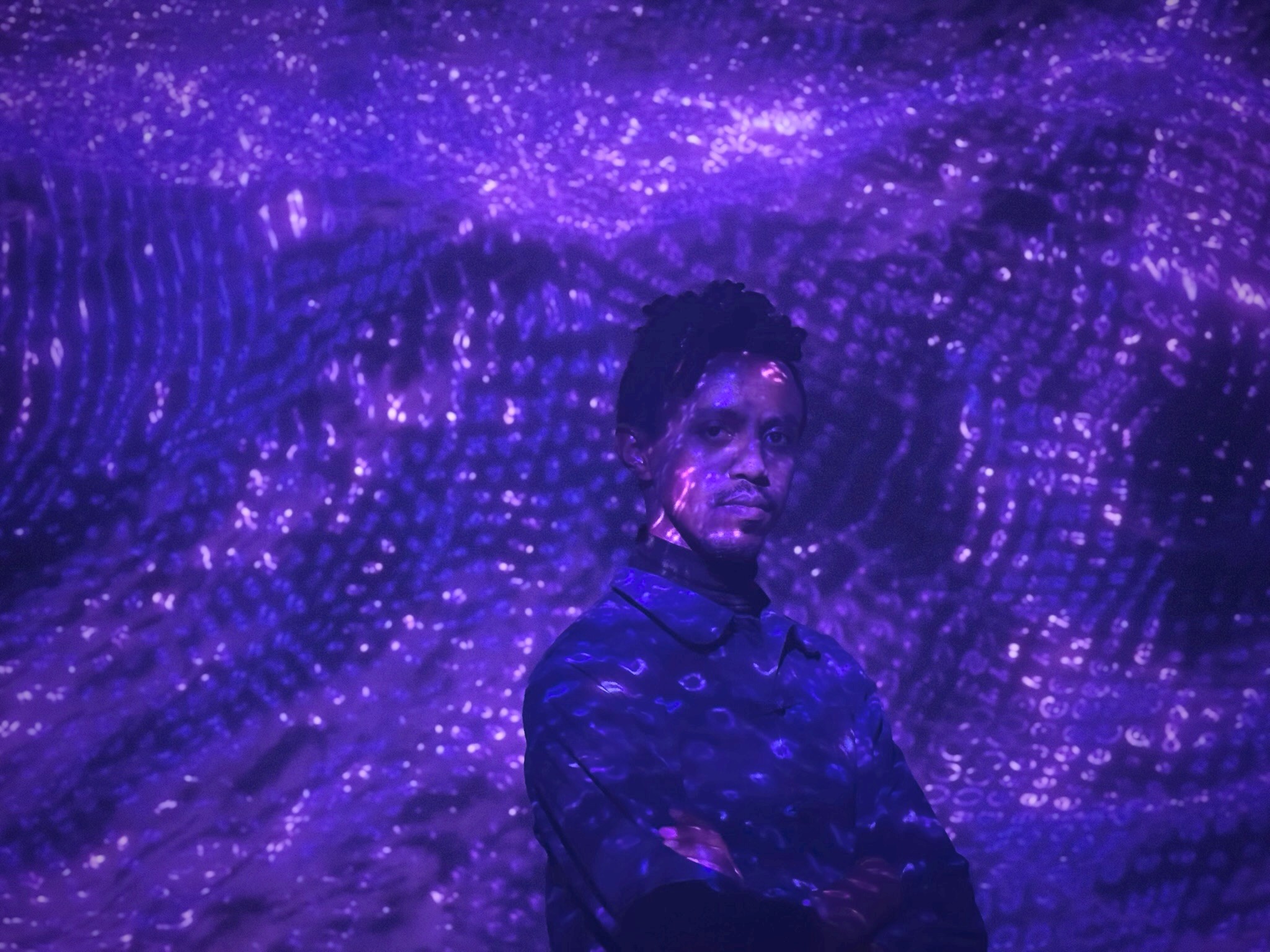 Portrait of Kidus Hailesilassie, of Ethiopian lineage. He stands before a swirling wave of indigo light, partially illuminated by projected patterns that ripple across his face and clothing. He is wearing a dark collared jacket, with arms folded at his chest. The photo dissolves the boundary between his presence and oceanic current, a luminous cosmic backdrop of shifting textures.