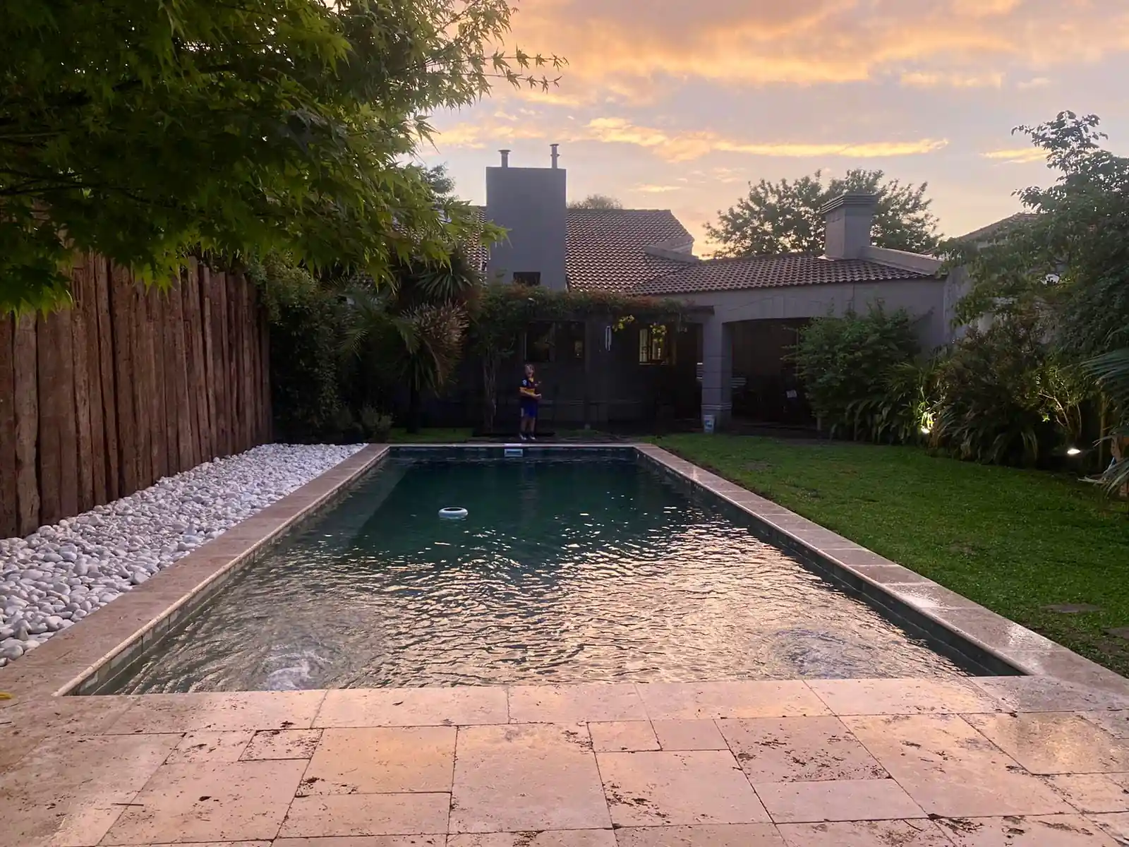 Vista al atardecer de una pileta con bordes de travertino, sector de piedras blancas decorativas y un amplio jardín de fondo.