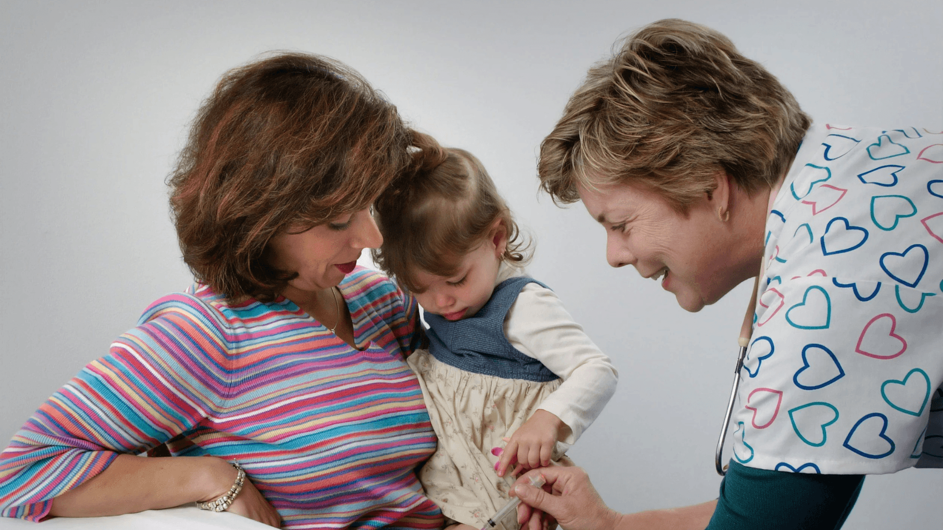 A healthcare professional smiling while showing a toddler a medical syringe as the child sits on a caregiver’s lap.