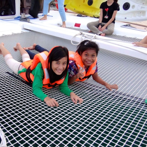两位微笑的女孩穿着救生衣躺在船的网上，其他人则坐在甲板上。