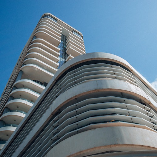 Tall, modern building with curved balconies and horizontal lines, viewed from below against a clear blue sky.