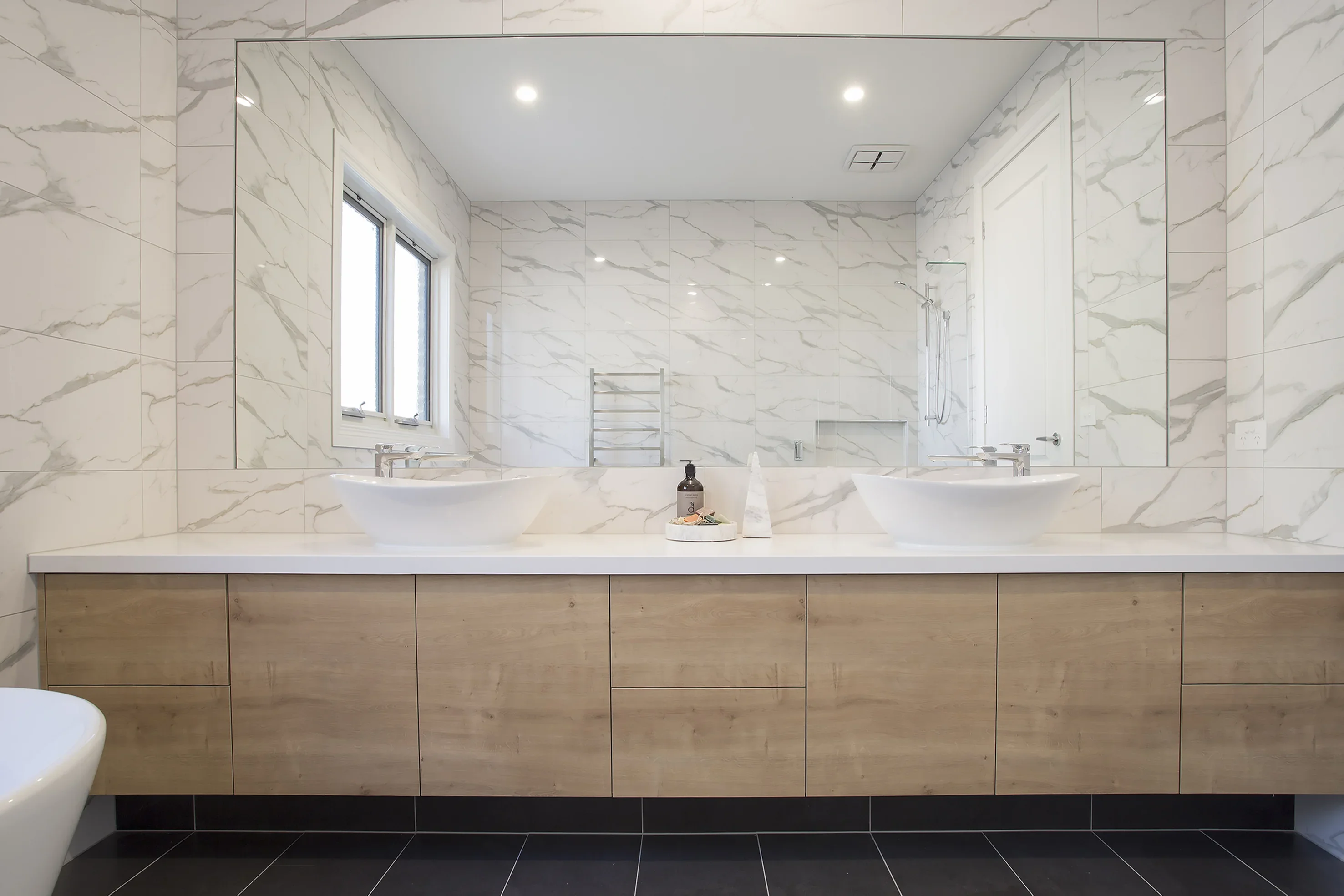 Bathroom with twin white vessel basins, a large frameless mirror, marble-look wall tiles, timber vanity and dark floor tiles