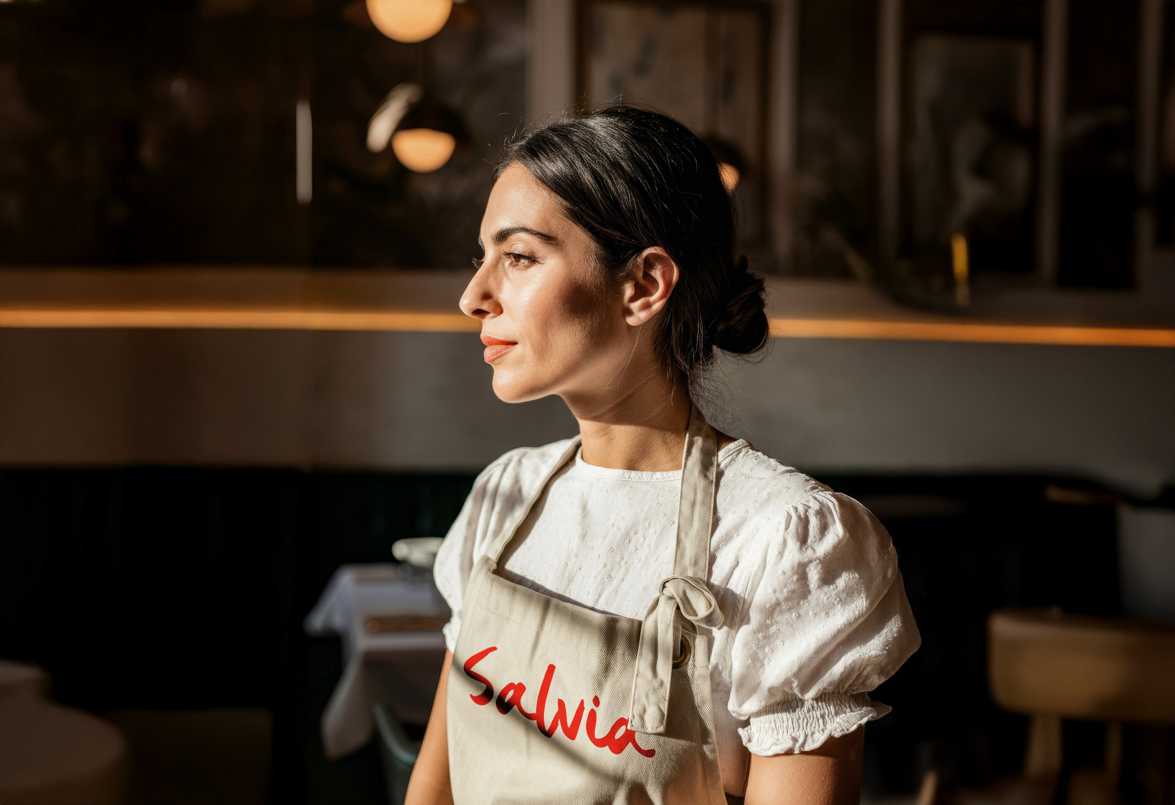 A woman with dark hair tied in a bun, wearing a white blouse and an apron with "Salvia" written on it, is gazing thoughtfully to the side in a dimly lit, elegant restaurant setting.