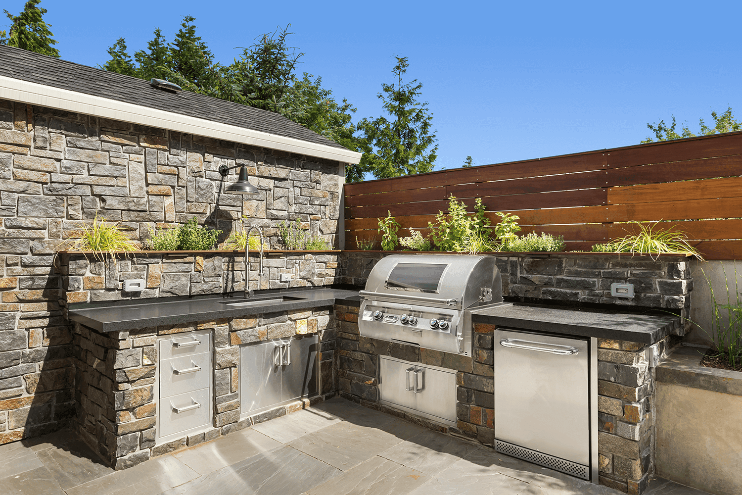 A built-in stainless steel grill set within a stone outdoor kitchen counter, overlooking a serene lake surrounded by vibrant autumn trees. The countertop is styled with lanterns, a fruit bowl, and waffle cones, creating a cozy outdoor dining vibe.