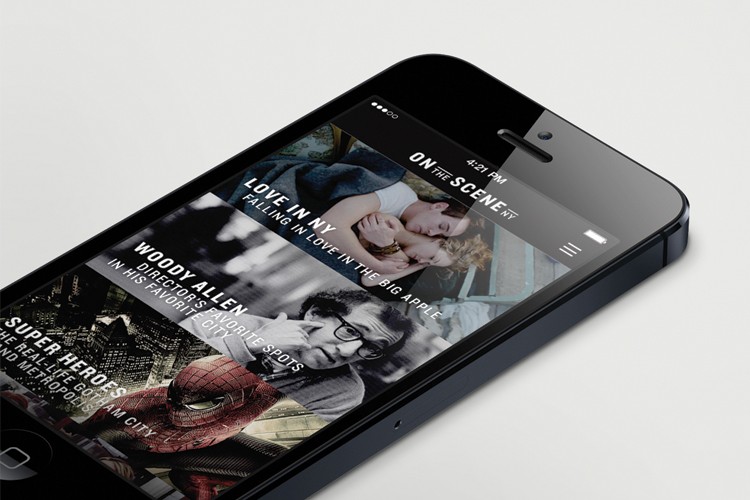 A black iPhone lies on a white surface, angled slightly to the right. The screen displays a movie app interface. The top bar shows "4:21 PM" and "ON THE SCENE NY." The main content area displays two movie posters: "LOVE IN NY" and "SUPER HEROES." Below them, a smaller poster for "WOODY ALLEN" shows a man with glasses and a red-and-black costume. The time displayed on the top bar is 4:21 PM.