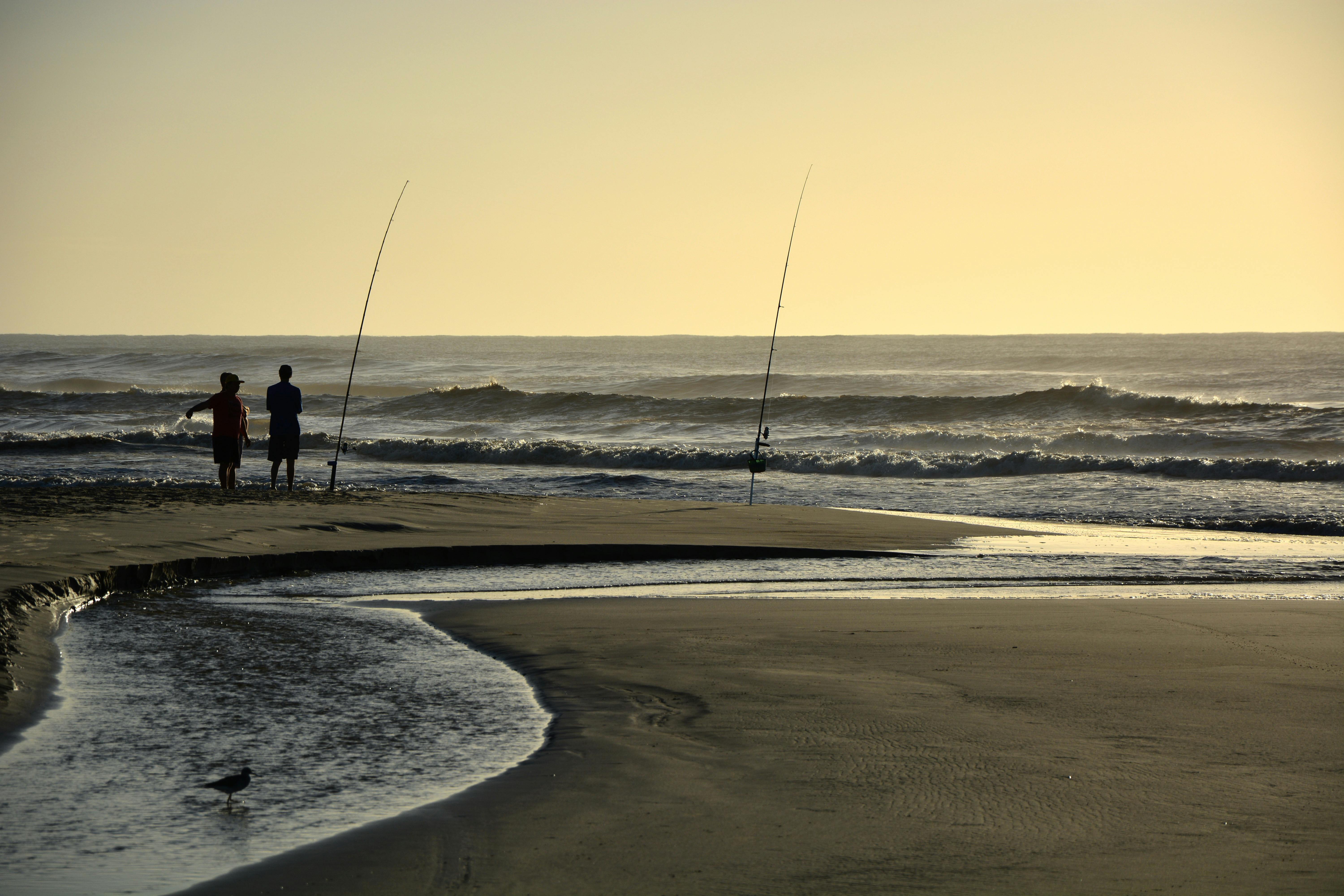 Two people stand by the water's edge on a beach at sunset, with gentle waves lapping at the shore.
