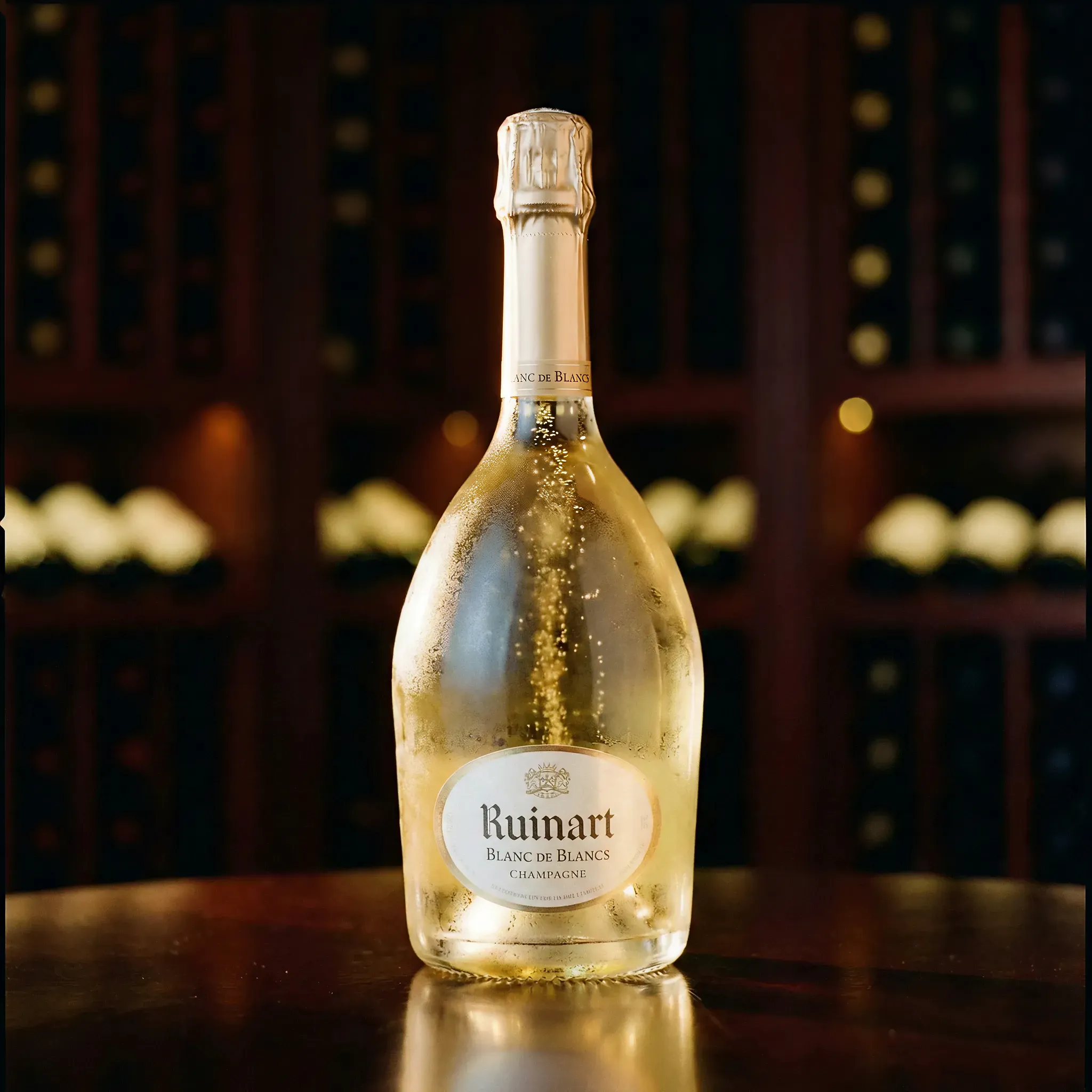 A chilled bottle of Ruinart Blanc de Blancs champagne sits on a wooden table, with a soft-focus background of wine racks. The mood is elegant and celebratory.