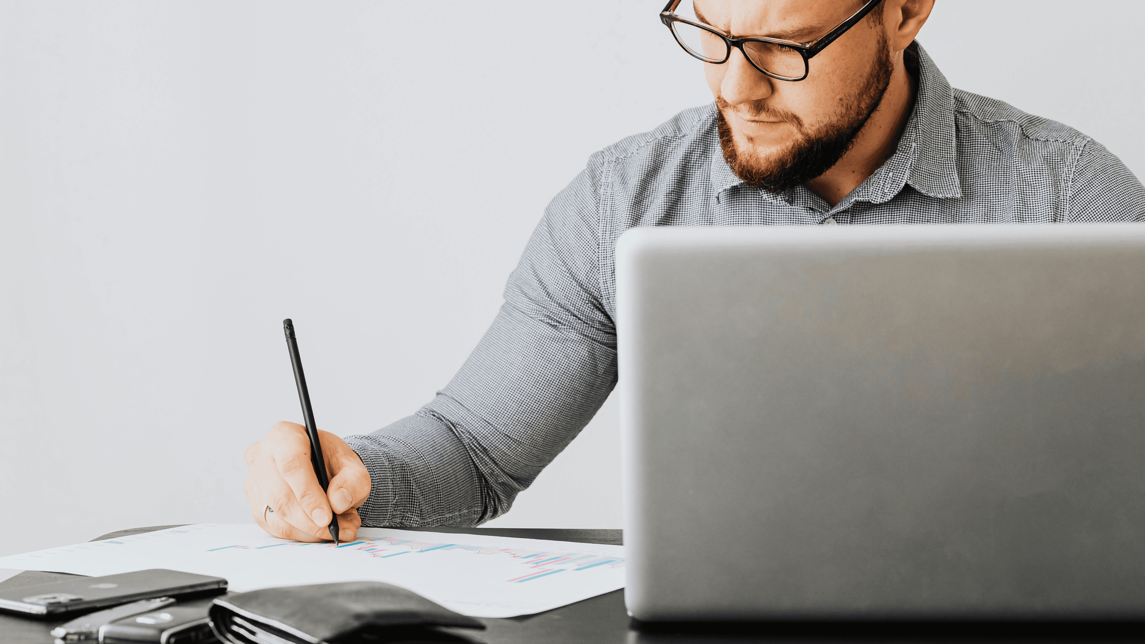 Homme travaillant à un bureau, prenant des notes sur papier à côté d’un ordinateur portable, avec des graphiques et des documents étalés, représentant un travail de planification ou d’analyse