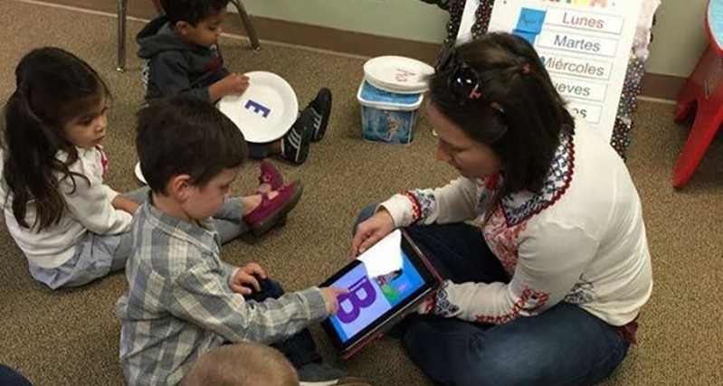 A photo of a teacher showing to a group of children the letters of the alphabet on a tablet.
