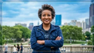 Actress and comedian Wanda Sykes stands confidently with arms crossed, smiling, against a backdrop of a cityscape and greenery.