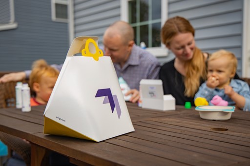 A family enjoys outdoor time at a wooden table with toys and packaged food, set against the backdrop of a gray-sided house.